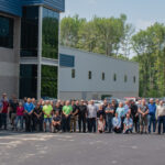 Group of SDC employees pose for a photo in front of their modern office building. The building features dark blue and gray facade and reflective glass windows.
