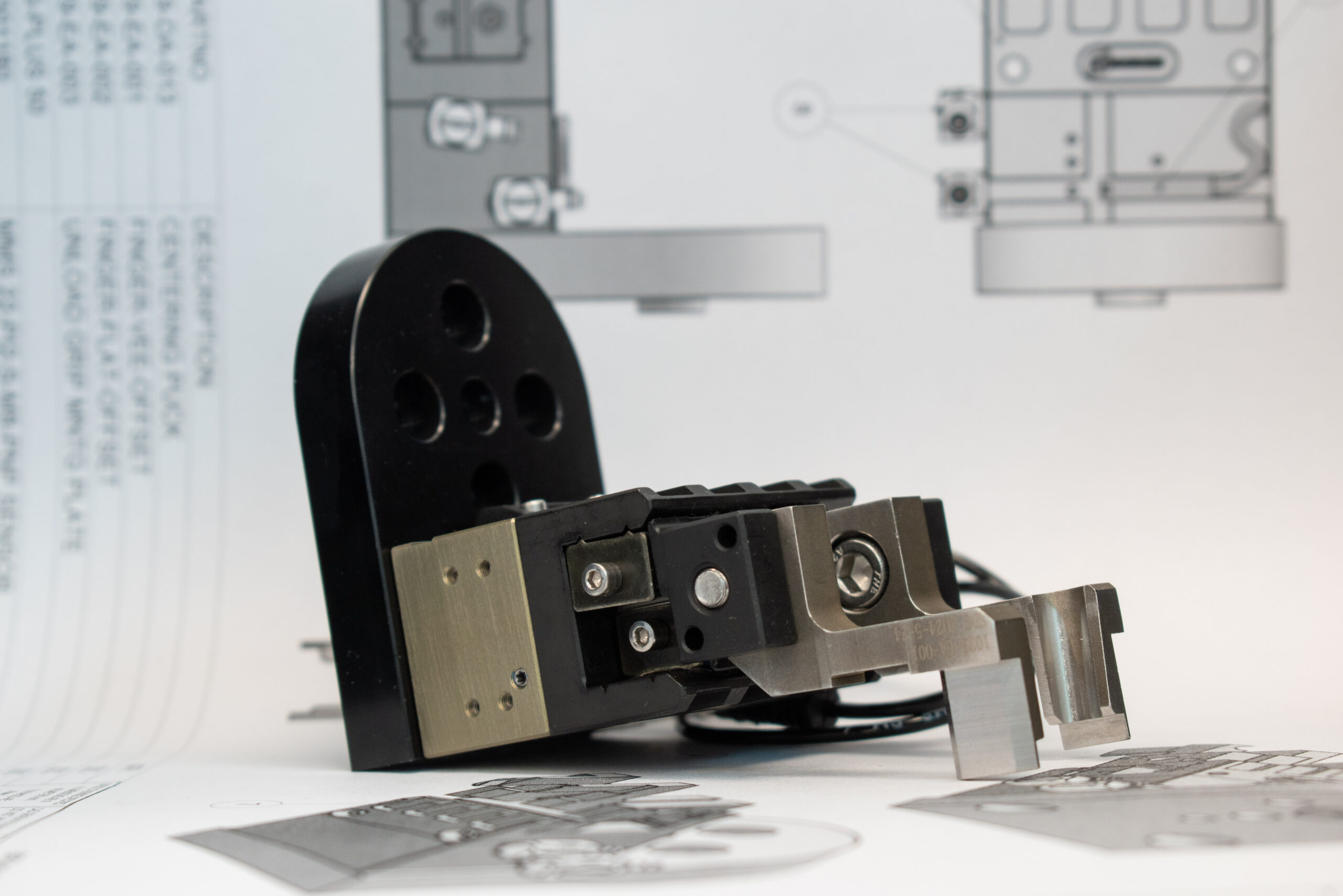 Close-up of a robotic arm component, featuring various metal parts in silver, black, and gold tones, set against a white background overlaid with technical engineering drawings.