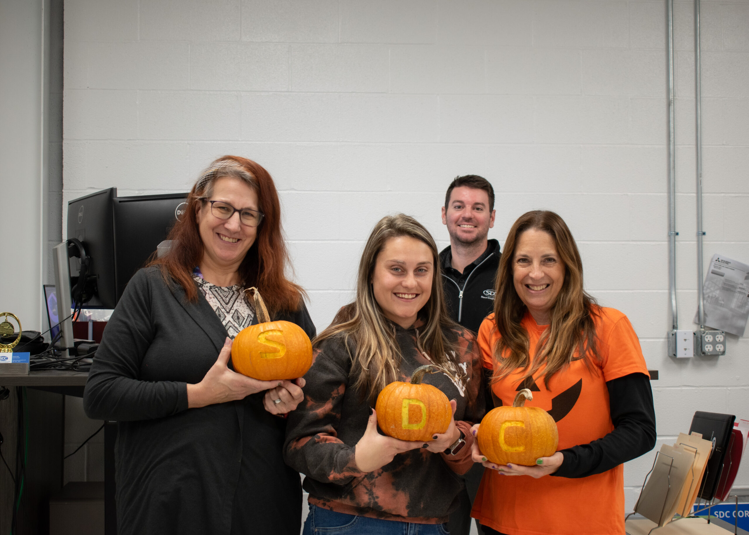 Team holding pumpkins carved with "SDC" letters, celebrating Halloween in an office environment.