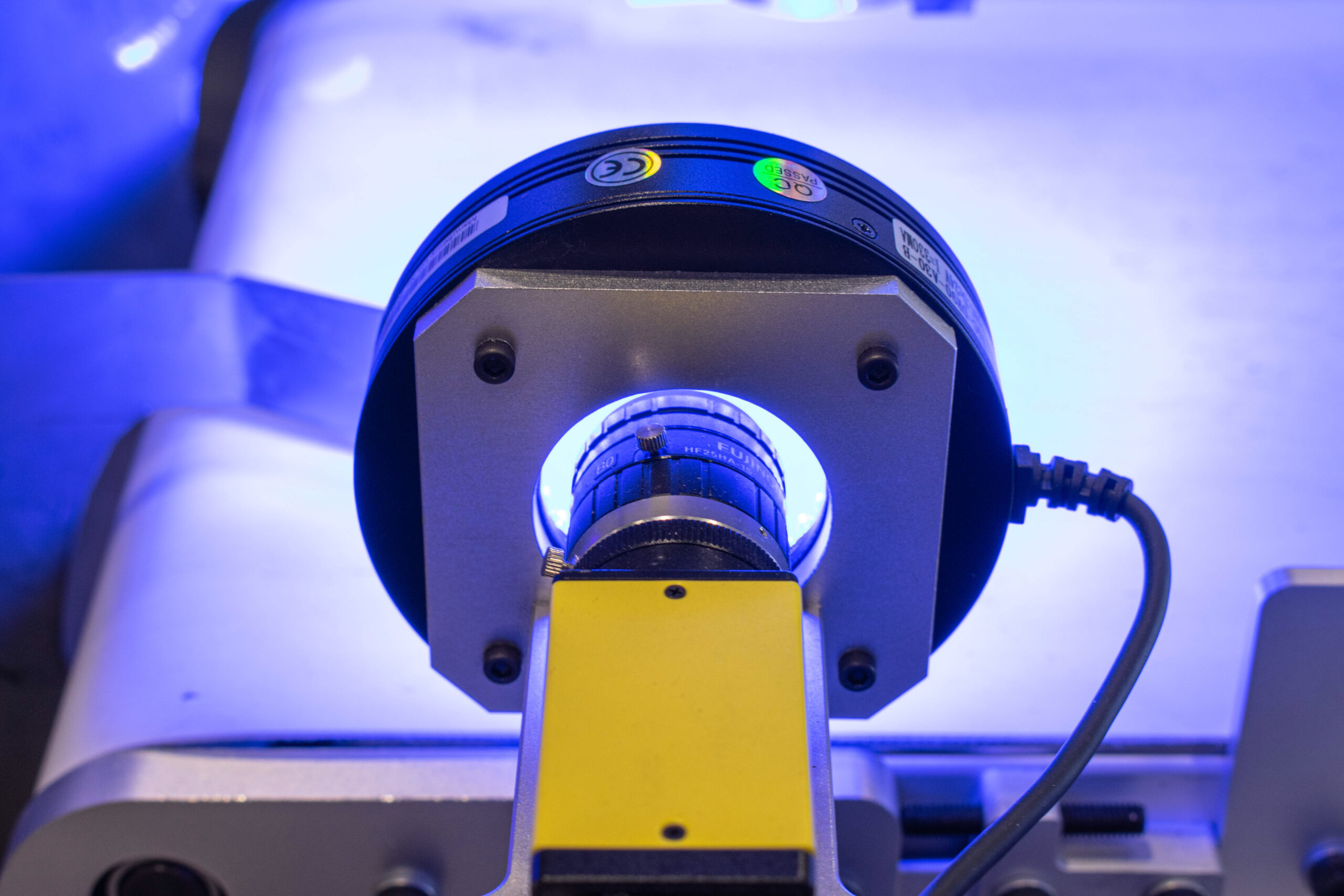 A close-up shot of an industrial inspection camera. The camera assembly includes a black cylindrical casing fixed to a silver-colored metal mounting plate with four screws.