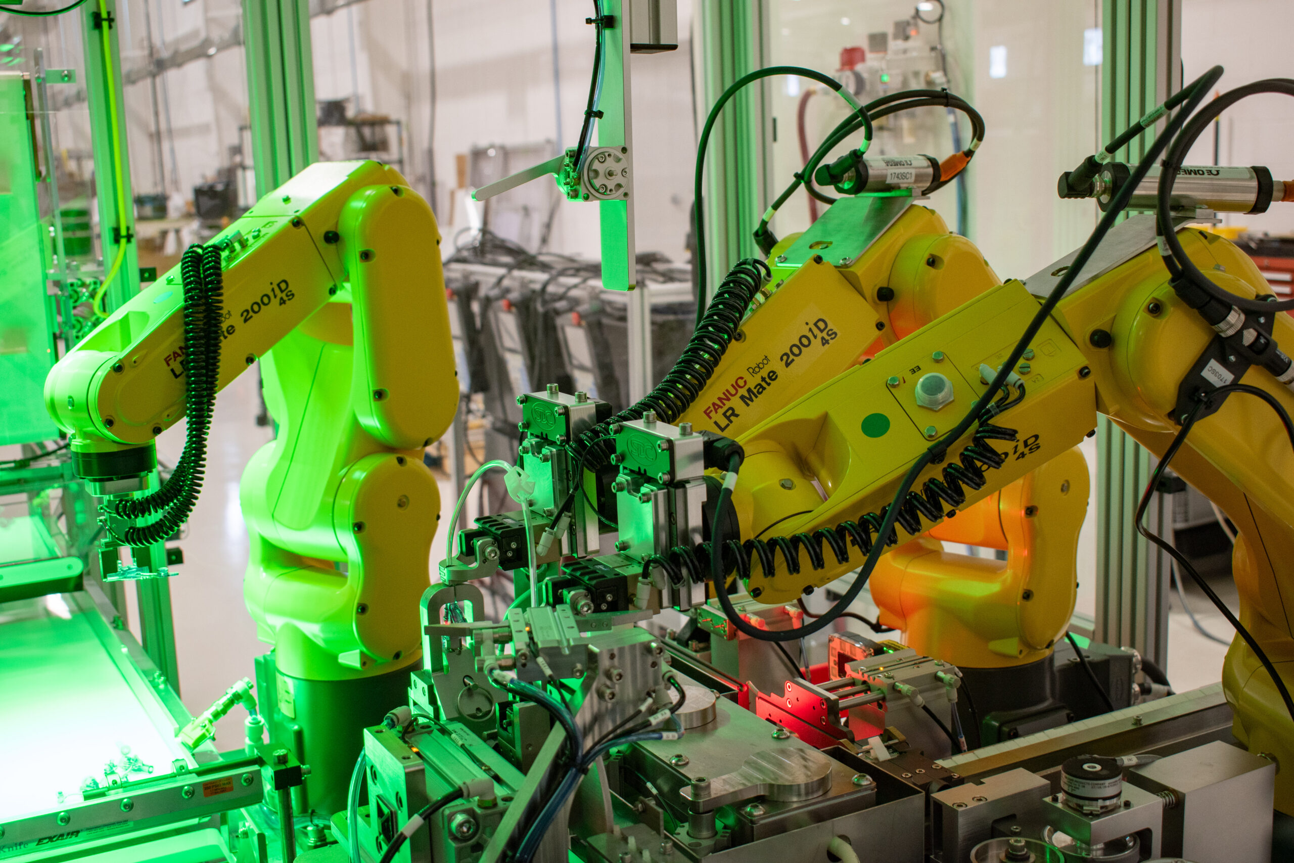 Two FANUC robots performing synchronous robotic bonding while a third FANUC robot is queued to load a part